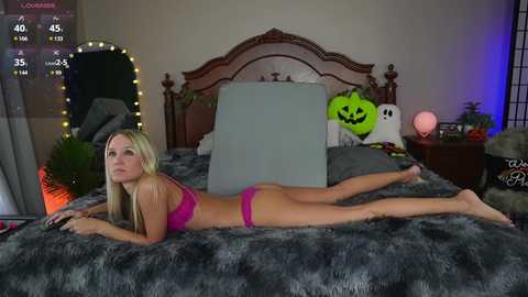 Media: Video of a blonde woman in pink lingerie lying on a bed with a grey fur blanket, surrounded by Halloween decorations and a TV displaying game stats.