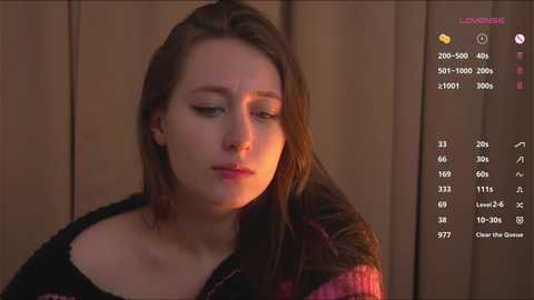 Media: Video of a young woman with light skin and straight brown hair, wearing a black and pink sweater, looking thoughtful against a wooden background.