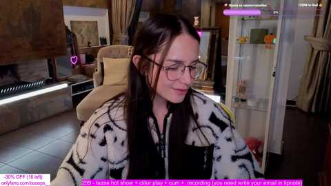 Media: Video of a young woman with long dark hair, wearing glasses, a black-and-white patterned jacket, sitting in a cozy living room with a fireplace, a glass cabinet, and plush seating.