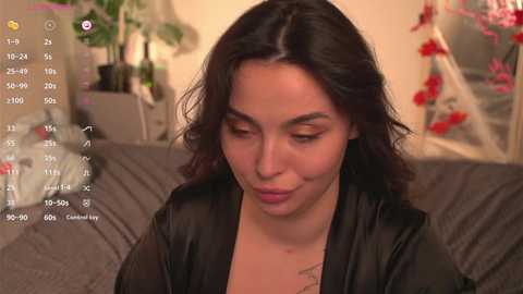 Media: Video of a Latina woman with dark hair, wearing a black satin robe, sitting on a gray couch. Background features a Christmas tree with red ornaments and a green plant.