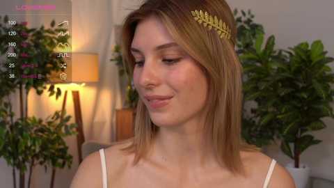 Media: Video of a fair-skinned woman with shoulder-length, light brown hair adorned with a golden laurel headband. She has a serene expression. Background includes potted plants and a lamp.