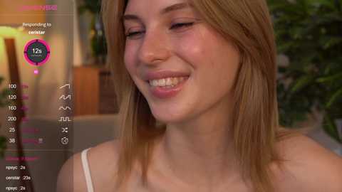 Media: Video of a smiling young woman with light skin and straight, shoulder-length blonde hair. She wears a white tank top, and the background shows a blurred, cozy indoor setting.