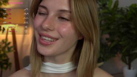 Media: Video of a smiling, fair-skinned young woman with straight, shoulder-length blonde hair, wearing a white choker necklace. Background features a potted plant and dimly lit room.