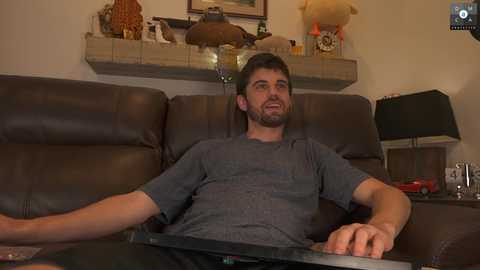 Media: Video of a smiling, light-skinned man with short brown hair, wearing a gray t-shirt, lounging on a brown leather couch in a cozy living room.