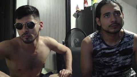 Media: Video of two shirtless men, one with short dark hair and sunglasses, and the other with long hair in a black and white patterned tank top, sitting in a dimly lit room with a fan and a refrigerator in the background.