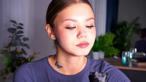 Media: Video of a fair-skinned, light-haired young woman with a small tattoo on her neck, wearing a blue shirt, applying makeup in a well-lit, indoor room with potted plants and a glass of water on a table.