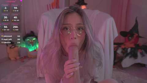 Media: Video of a young Asian woman with long blonde hair, wearing glasses, performing oral sex on a large, erect penis. Background includes a plush toy, Halloween decorations, and a gaming setup.