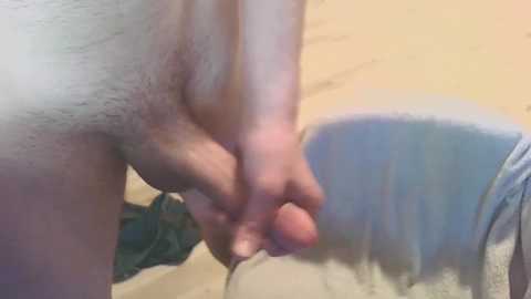 Media: A close-up video of a nude male, showing his partially erect penis and testicles. He is standing against a beige wall, with a blue towel draped over a chair in the background. The image is candid and intimate, focusing on the genital area.