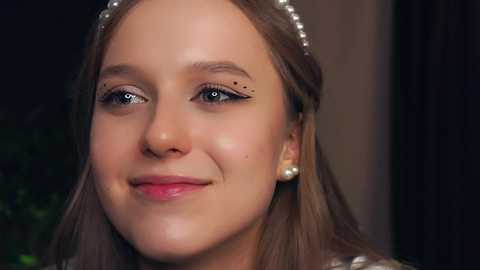 Media: Video of a young girl with fair skin, light brown hair, and a delicate smile. She wears a pearl headband and pearl earrings. Background is blurred, with a hint of green foliage.