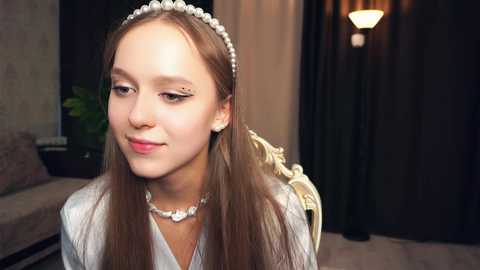 Media: Video of a young, fair-skinned woman with long brown hair, wearing a pearl headband, white dress, and pearl necklace, seated indoors with a beige sofa, green plant, and dark curtains in the background.