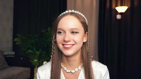 Media: Video of a young, smiling girl with light brown hair styled in two braids, wearing a white satin dress, pearl necklace, and headband, against a dark curtain backdrop with a lit lamp.