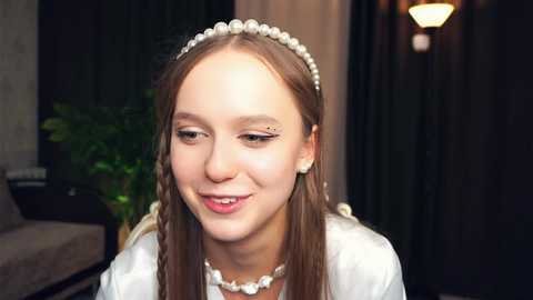 Media: Video of a young Caucasian woman with long brown hair, wearing a pearl headband and pearl necklace, smiling. Background shows a dark curtain and a potted plant.