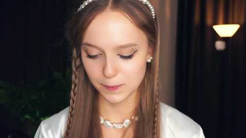 Media: Video of a young girl with long, straight brown hair, styled in two braids. She wears a white dress and a silver headband with pearls. The background is blurred, featuring warm lighting and dark curtains.