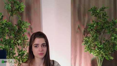 Media: Video of a young woman with long, straight brown hair, standing in front of two tall, green potted plants against a pink and beige wall. She wears a casual, dark-colored top.