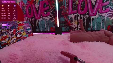 Media: Video of a vibrant, pink-fur-covered room with a plush rug. Walls are adorned with \"LOVE\" graffiti, neon lights, and colorful artwork. A large, realistic-looking dildo rests on the rug.