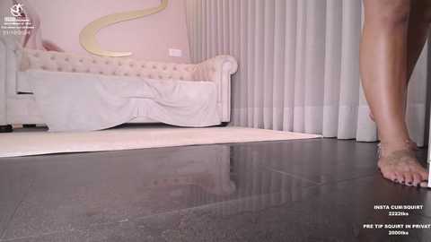 Media: Video of a woman's barefoot, tan legs standing on a dark marble floor in a minimalist bedroom with a tufted, white upholstered bed and sheer curtains.