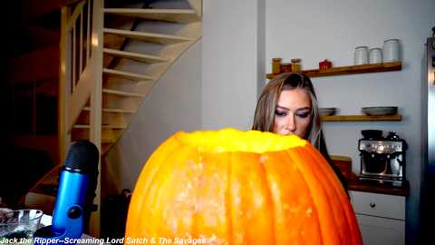 Media: Video of a woman with long brown hair, wearing a black top, carving a large orange pumpkin in a cozy kitchen with wooden shelves, a coffee maker, and a spiral staircase.