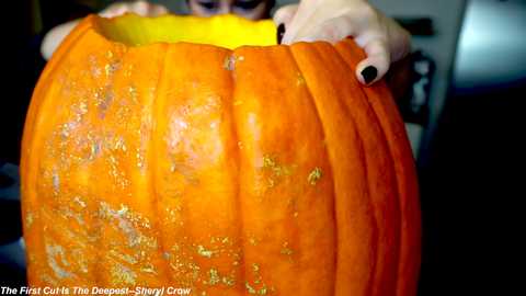 Media: Video of a person holding a carved, orange pumpkin with a gaping mouth, wearing black nail polish. Background is dark and blurry, focusing attention on the pumpkin.