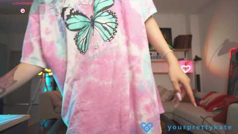 Media: A video of a young woman wearing a tie-dye T-shirt with a large butterfly design, standing in a modern living room with a TV, couch, and colorful lighting.