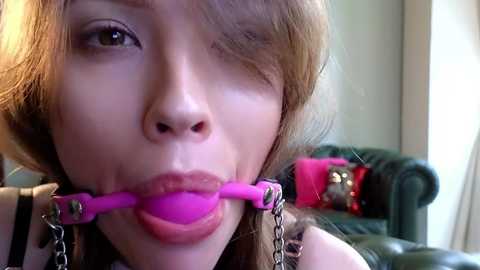 Media: Video of a young woman with fair skin, brown hair, and light makeup, wearing a pink ball gag and black leather collar with chains, in a living room with a green couch and pink pillow.