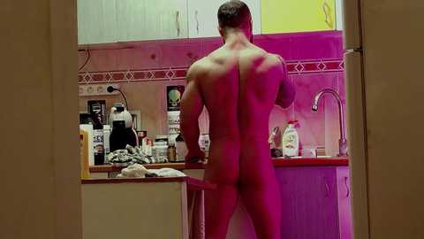 Media: Video of a naked man with a muscular build, viewed from behind, standing at a kitchen counter. The kitchen has pink and yellow cabinets, a sink, and various food items.