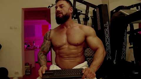 Media: Video of a muscular, shirtless man with a full beard, tattoos, and short dark hair, sitting in a home gym with a keyboard, surrounded by workout equipment and a pink-lit background.