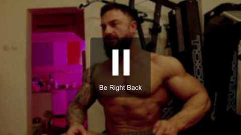 Media: Video of a shirtless, muscular man with tattoos and short dark hair, working out on gym equipment. Background includes a doorway with purple lighting and gym equipment. Text overlay: \"Be Right Back.\