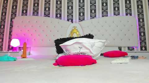 Media: Video of a bed with white tufted headboard, black and white striped wallpaper, pink and orange pillows, white and black decorative cushions, and a small pink vibrator.