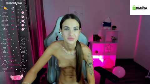 Media: Video of a topless, slim, light-skinned woman with long brown hair, sitting in a pink gaming chair, surrounded by gaming accessories and a pink background.