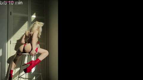 Media: Video of a blonde woman in red lingerie and boots, sitting on a chair, back to the camera, in a dimly lit room with white shutters.