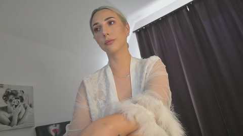 Media: Video of a woman with platinum blonde hair, wearing a sheer white lace robe, standing in a modern bedroom with dark curtains and a black and white framed photo on the wall.