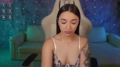 Media: Video of a young woman with long black hair, wearing a floral-patterned top, seated in a beige armchair against a blue, starry-patterned wall.