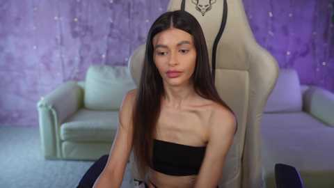 Media: Video of a slender, long-haired, Asian woman with small breasts, wearing a black strapless top, seated in a beige gaming chair. Background features a purple textured wall and a white sofa.