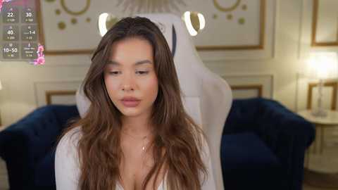 Media: Video of a young woman with long, wavy brown hair and light skin, wearing a white robe, sitting in a luxurious, dimly-lit room with a navy blue velvet sofa, a white lamp, and a framed picture in the background.