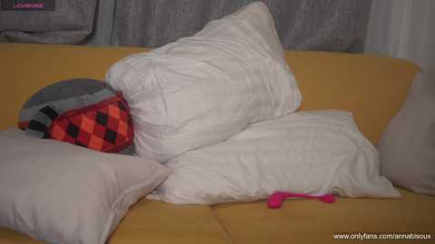 Media: Video of a cozy living room scene featuring a mustard-yellow sofa, a gray curtain, a red and black checkered pillow, and white pillows, with a pink vibrator lying on the sofa.