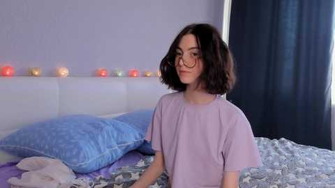 Media: Video of a young woman with shoulder-length, wavy dark hair, wearing glasses and a light purple t-shirt, sitting on a bed with blue pillows and a floral-patterned blanket, in a softly lit room with string lights and a dark curtain in the background.