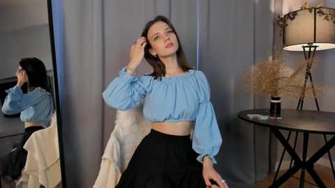 Media: Video of a Caucasian woman with straight, shoulder-length dark hair, wearing a blue blouse and black skirt, standing in front of a mirror and a round table with a lamp.