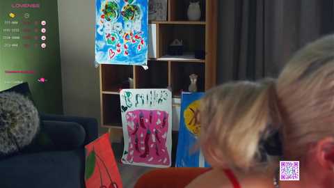 Media: Video of a cozy living room with a woman in a red top, focusing on colorful children's art pinned on a wooden shelf.