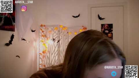 Media: A video of a woman with long brown hair, wearing a brown jacket, inside a Halloween-themed room with bat decorations, illuminated by fairy lights.