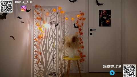 Media: Video of a Halloween-themed room with a white folding screen adorned with orange and black spider webs and bats, a small yellow stool, and a vase with fake flowers. A framed photo of a smiling woman is on the door.