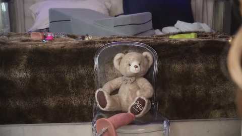 Media: Video of a vintage teddy bear in a clear acrylic chair on a fur rug, surrounded by a cluttered room with a bed, pillows, and assorted objects.