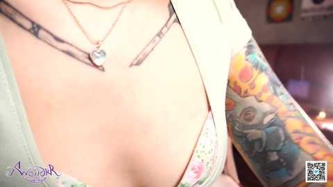 Media: Video of a woman with fair skin, large breasts, and a floral-patterned bra, wearing a white cardigan, showcasing colorful tattoos on her left arm and neck.