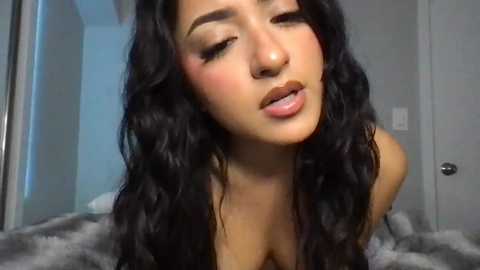 Media: A video of a young woman with long, wavy black hair, light brown skin, and full lips, wearing makeup, leaning forward with a sultry expression. She's indoors, on a bed with gray blankets.