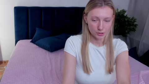 Media: A video of a blonde Caucasian woman with straight hair, wearing a white T-shirt, sitting on a bed with a dark headboard and pink quilt.