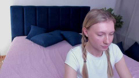 Media: Video of a young Caucasian woman with light skin and blonde hair in pigtails, wearing a white t-shirt, sitting on a bed with a pink quilt and dark blue pillows.