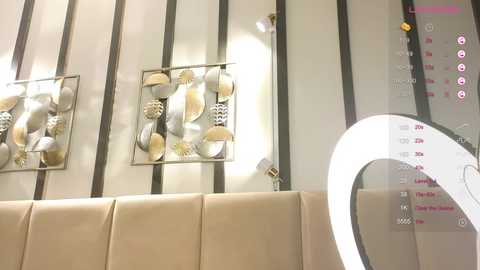 Media: Video of a modern, beige upholstered wall with three decorative, metallic leaf-shaped wall sculptures. A large circular mirror with a bright, white lightbulb is mounted on the right, reflecting the room's ambiance.