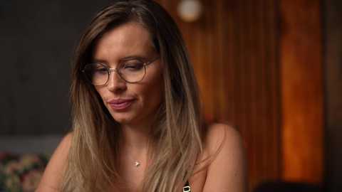 Media: Video of a fair-skinned woman with long, straight, blonde hair, wearing glasses and a black top, smiling softly. Background features a warm, wooden door and dimly lit room.