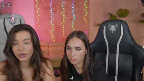 Media: Video of two young women with long hair, one topless, seated in black gaming chairs, in a colorful, festive room with multicolored string lights.