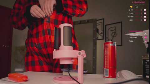 Media: Video of a person in a red and black plaid shirt using a pink, handheld hairdryer on a countertop cluttered with a red can, a bottle, and a phone.