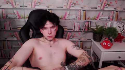 Media: Video of a shirtless, fair-skinned, young man with messy black hair, multiple tattoos, and a neutral expression, seated in a black gaming chair. Background features a bookshelf filled with colorful books and a small desk with a red toy, fern, and clock.